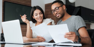 Photo of cheerful loving young couple using laptop and analyzing their finances with documents. Look at papers.